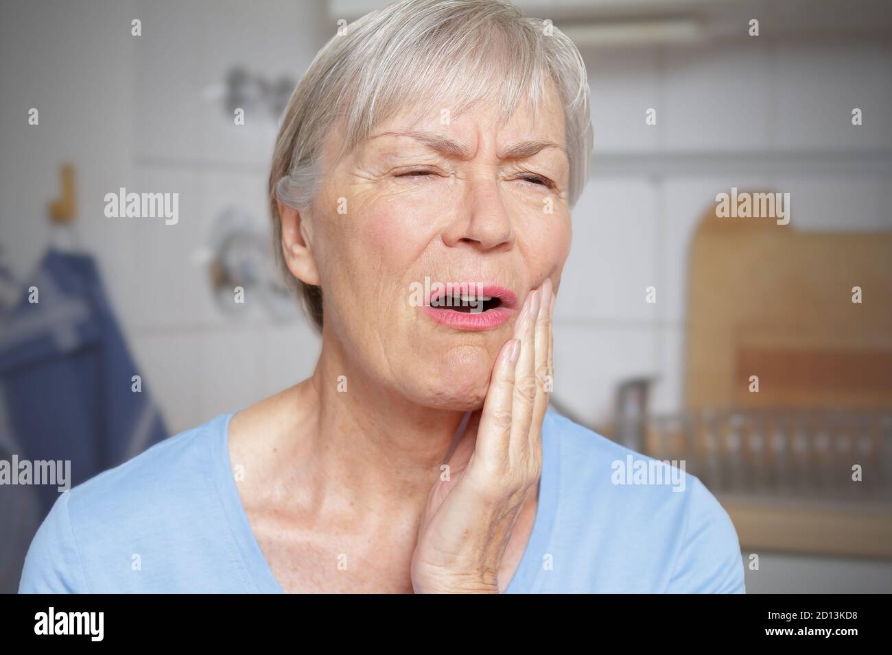 Acute toothache: elderly woman in her kitchen with a hand at her painful cheek. Stock Photo