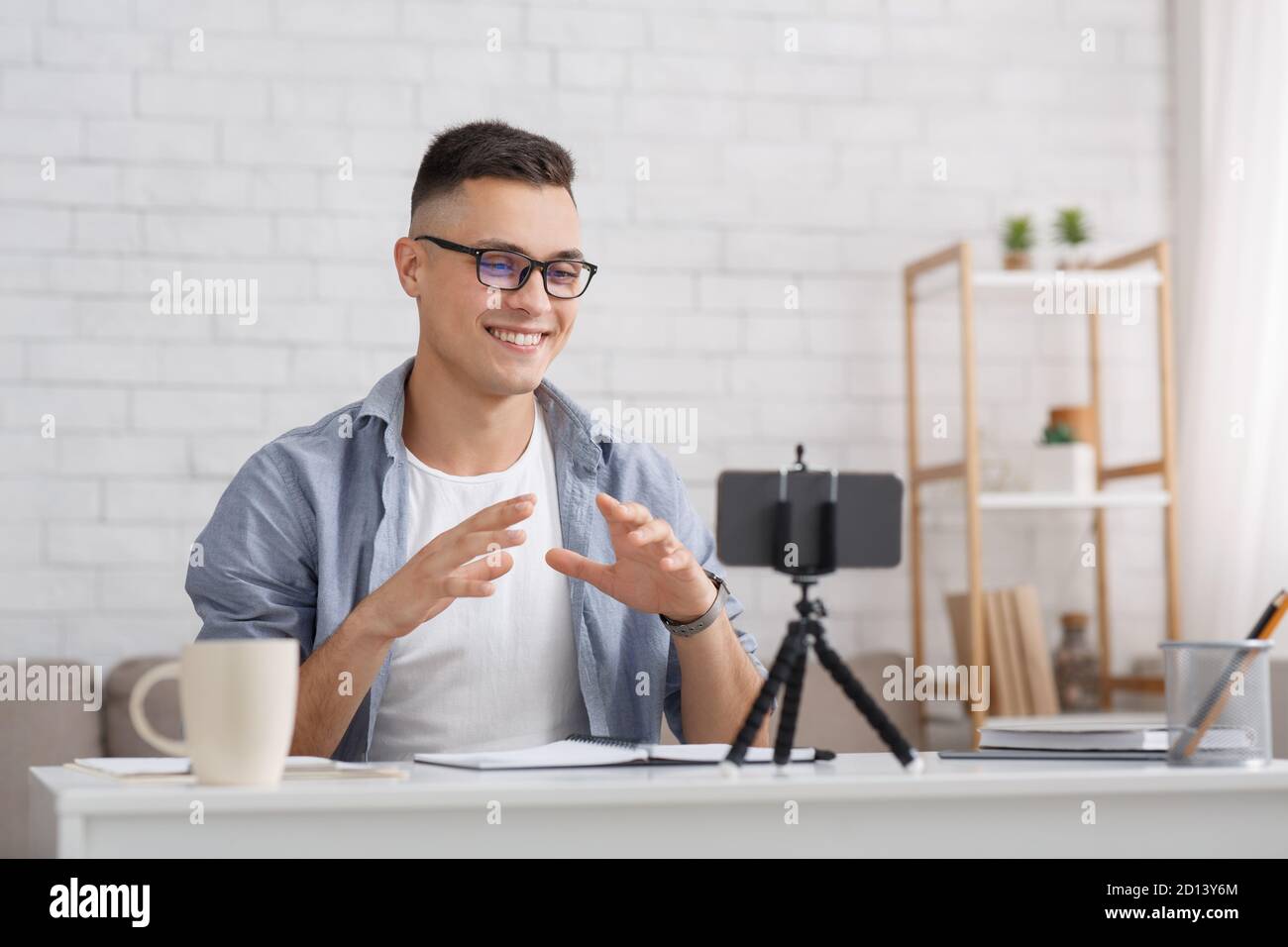 Modern devices for hobby or work at home. Smiling guy in glasses makes podcast, watch at webcam on tripod at living room during coronavirus epidemic Stock Photo