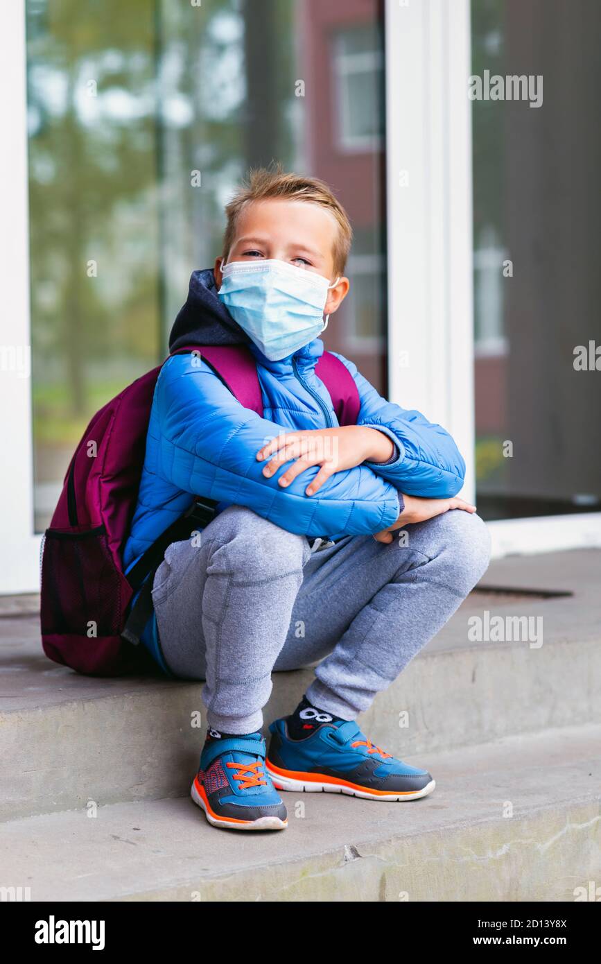 Full lenght Sad schoolkid boy child kid wearing face mask resting outdoors. Bored little student sitting down on the stairs and look at camera Stock Photo