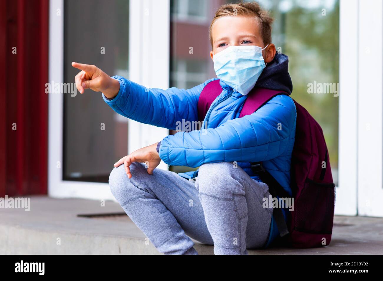 Sad schoolkid boy child kid resting outdoors. Bored little student look aside He wears medical mask pointing with his finger somewhere Stock Photo