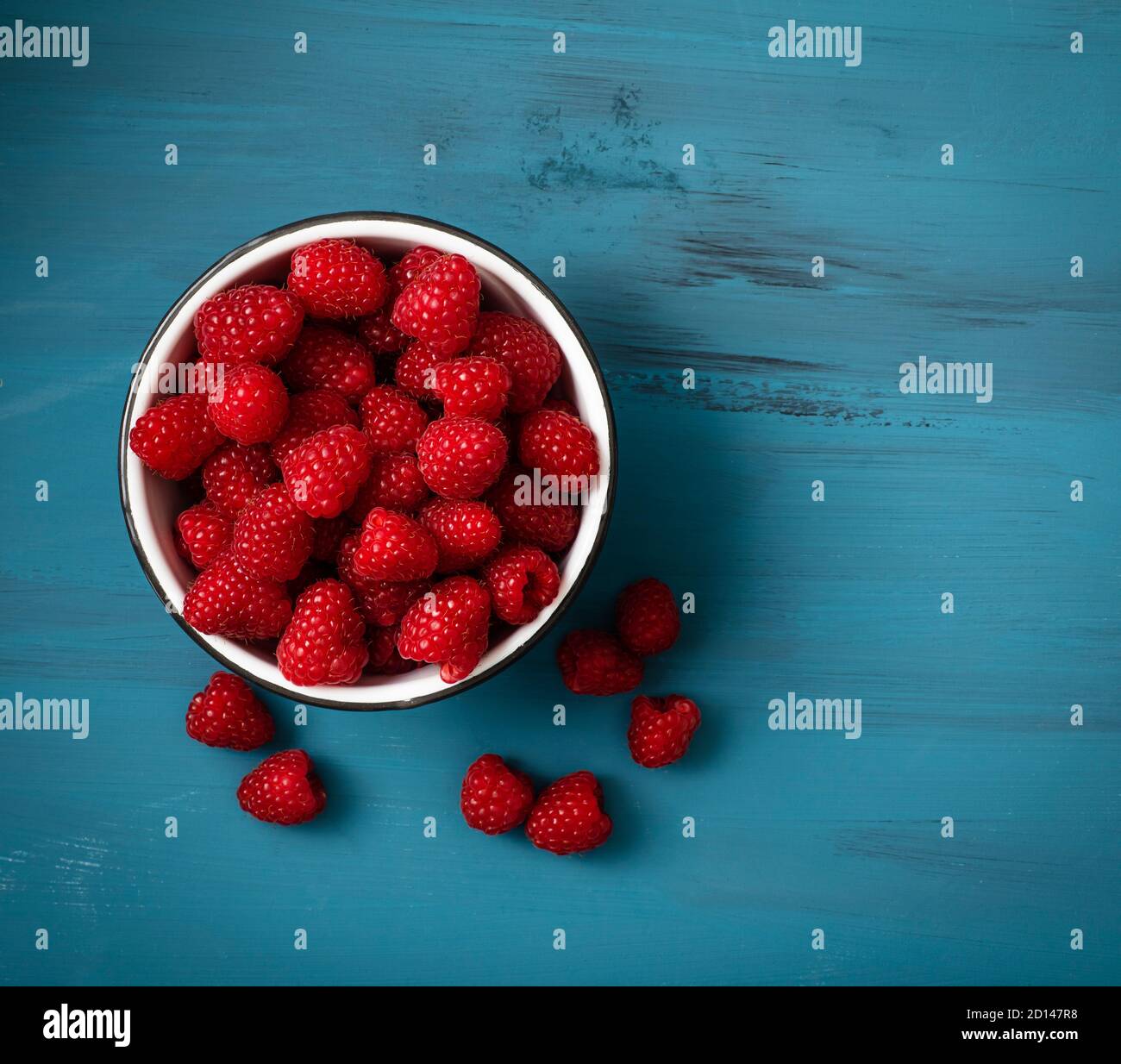Lots of raspberries in a bowl placed on rustic, colored background. Some berries are placed on the surface outsiden the bowl. Picture is shot from abo Stock Photo