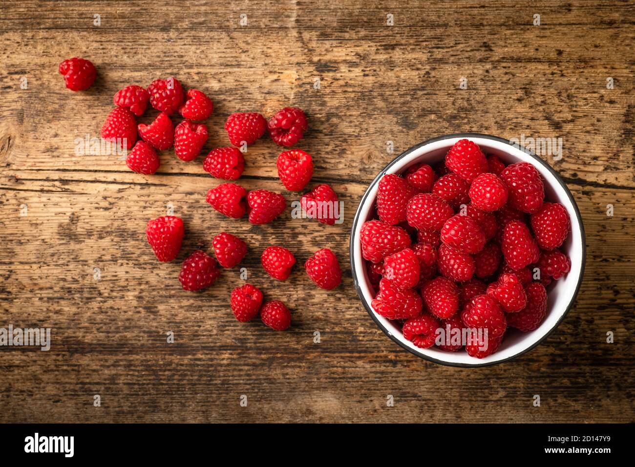 Lots of raspberries in a bowl placed on rustic, wooden background. Berries are placed on the surface outsiden the bowl. Picture is shot from above. Stock Photo