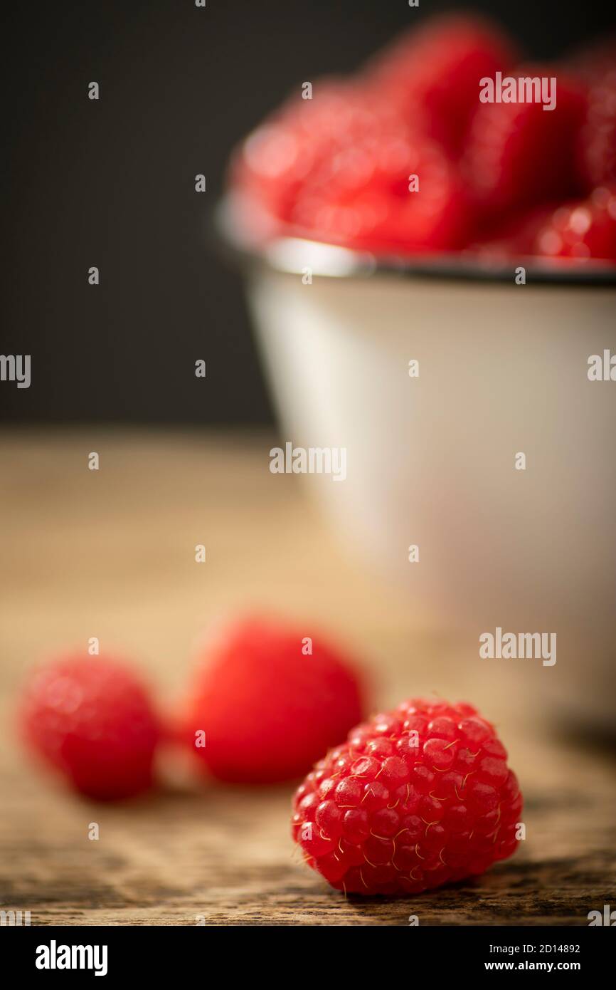 Closeup shot of a raspberry on a wooden table. Behind is a bowl of raspberries. against a dark grey background.  Shallow depth of field Stock Photo
