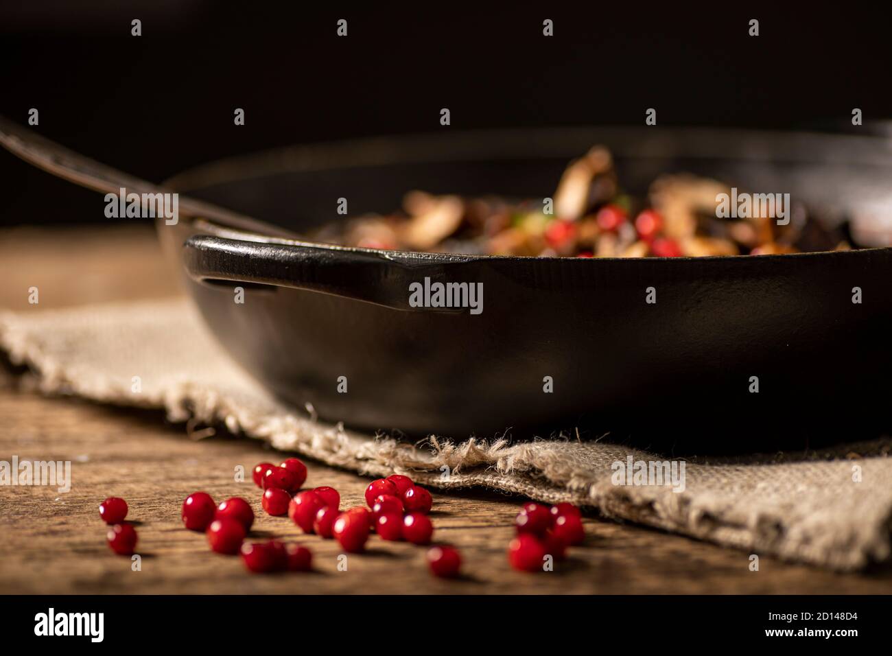 Bunch of lingonberries placed on a rustic wooden table. Black pot with food in background. Shallow depth of field. Stock Photo