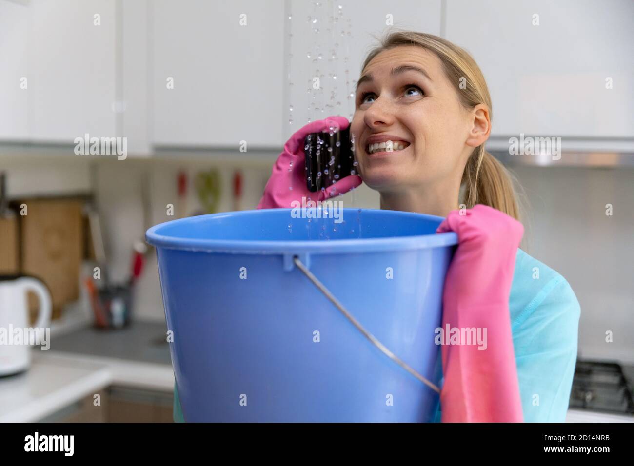 apartment flooded by upstairs - worried woman calling repair service while water leaking from ceiling in bucket Stock Photo