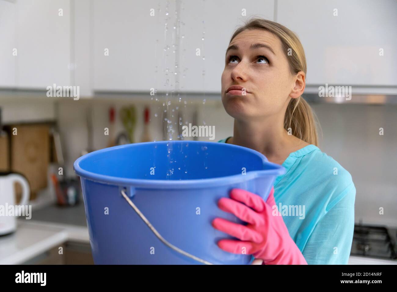 home flooded by upstairs or roof damage - woman holding bucket while water leaking from ceiling in kitchen Stock Photo