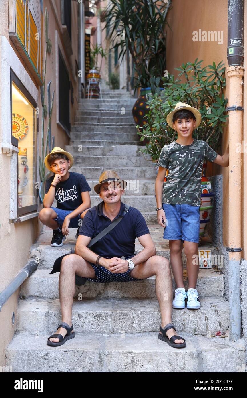 Italy, Sicily, Taormina, man and his two sons wearing straw hats sitting on on the steps of a sloping alleyway overlooking the corso umberto Stock Photo