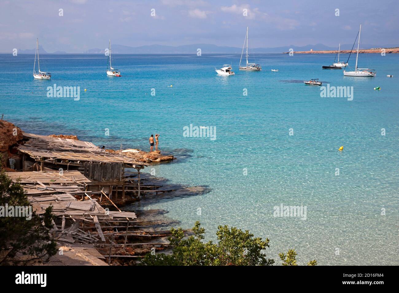 Spain, Balearic islands, formentera, Cala Saona, couple in swimming costume at the foot of of boat sheds watching sailboats mooring in turquoise water Stock Photo
