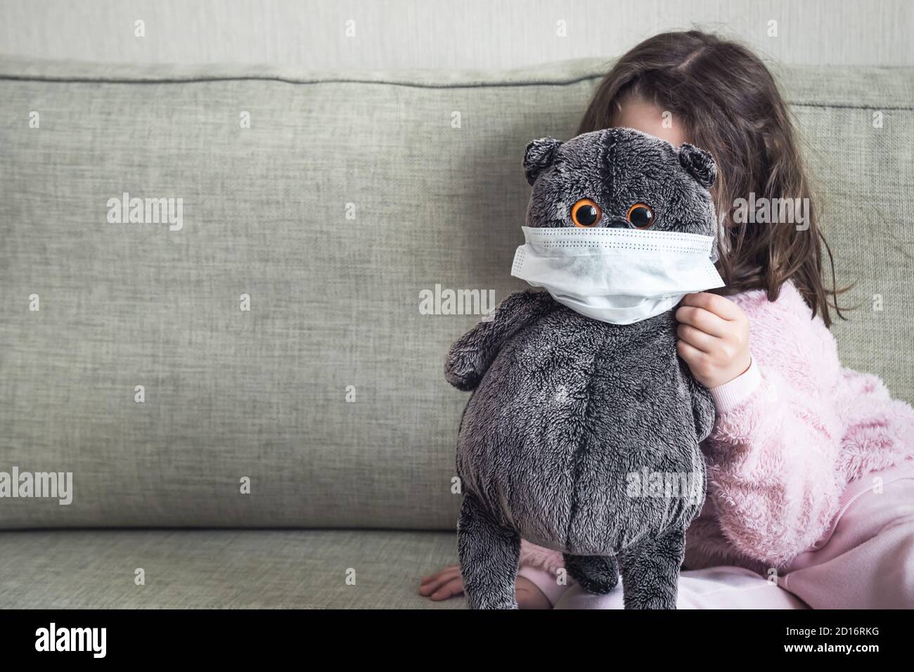 Kid playing with soft toy on couch during COVID-19 coronavirus pandemic, background for text. Playful child hides behind masked cat at home due to qua Stock Photo