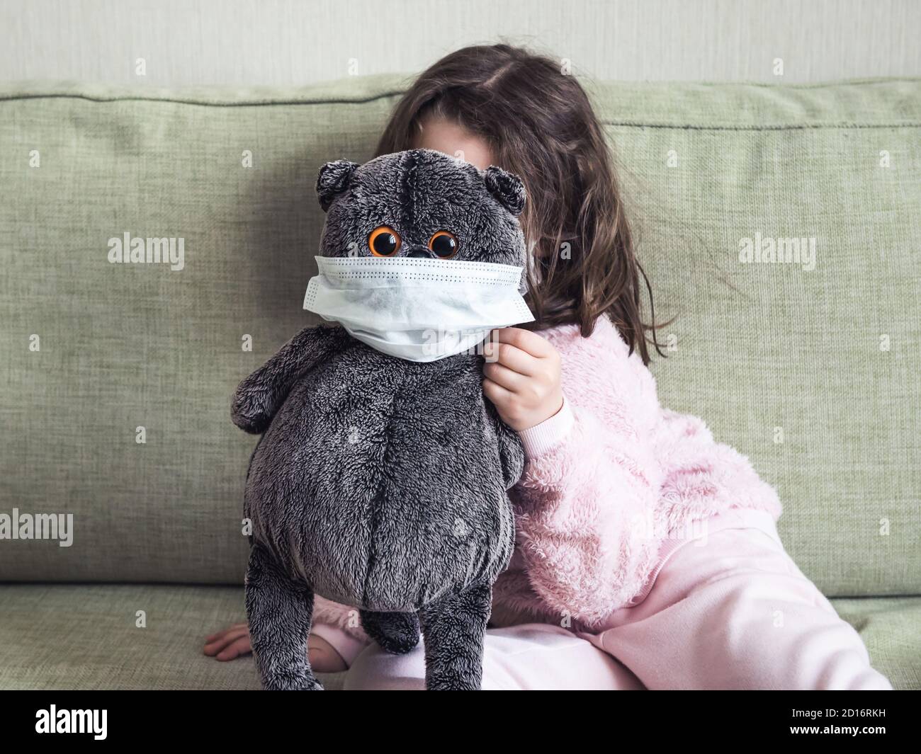 Kid playing with soft toy during COVID-19 coronavirus pandemic, funny playful little girl hides behind masked cat indoor due to quarantine. Concept of Stock Photo