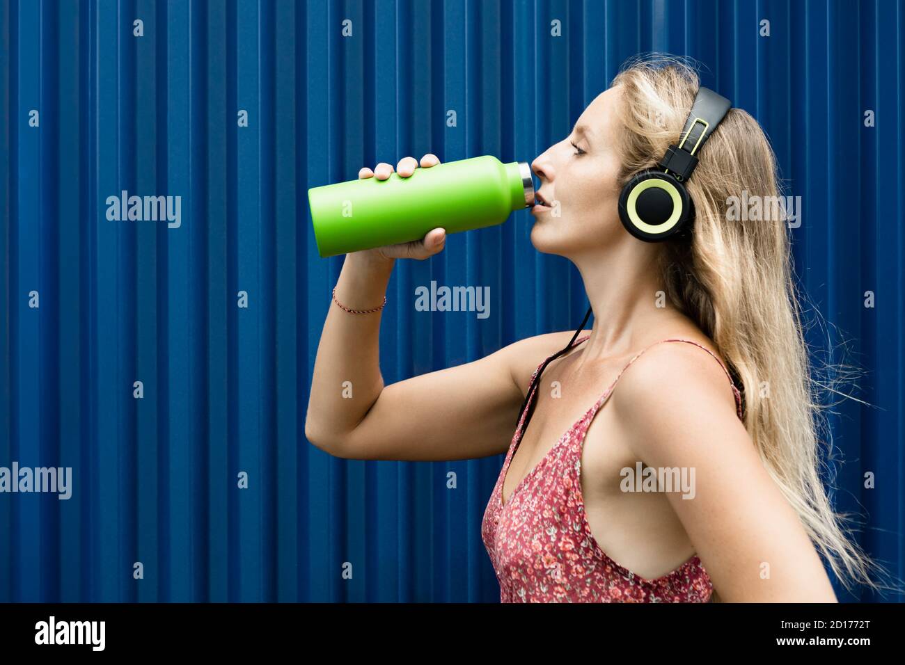 Young blond woman on blue background drink fresh spring water from green reusable bottle. Healthy lifestyle in summer city. Stock Photo