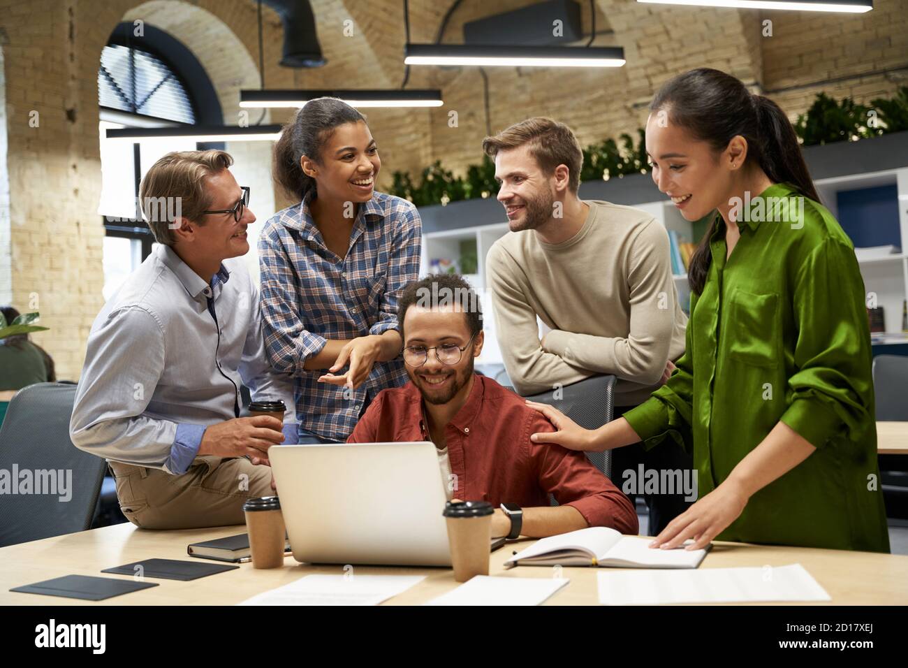 Sucssesful multiracial team analyzing project results. Group of young happy business people looking at laptop screen, communocating and discussing work while standing together in the modern office Stock Photo