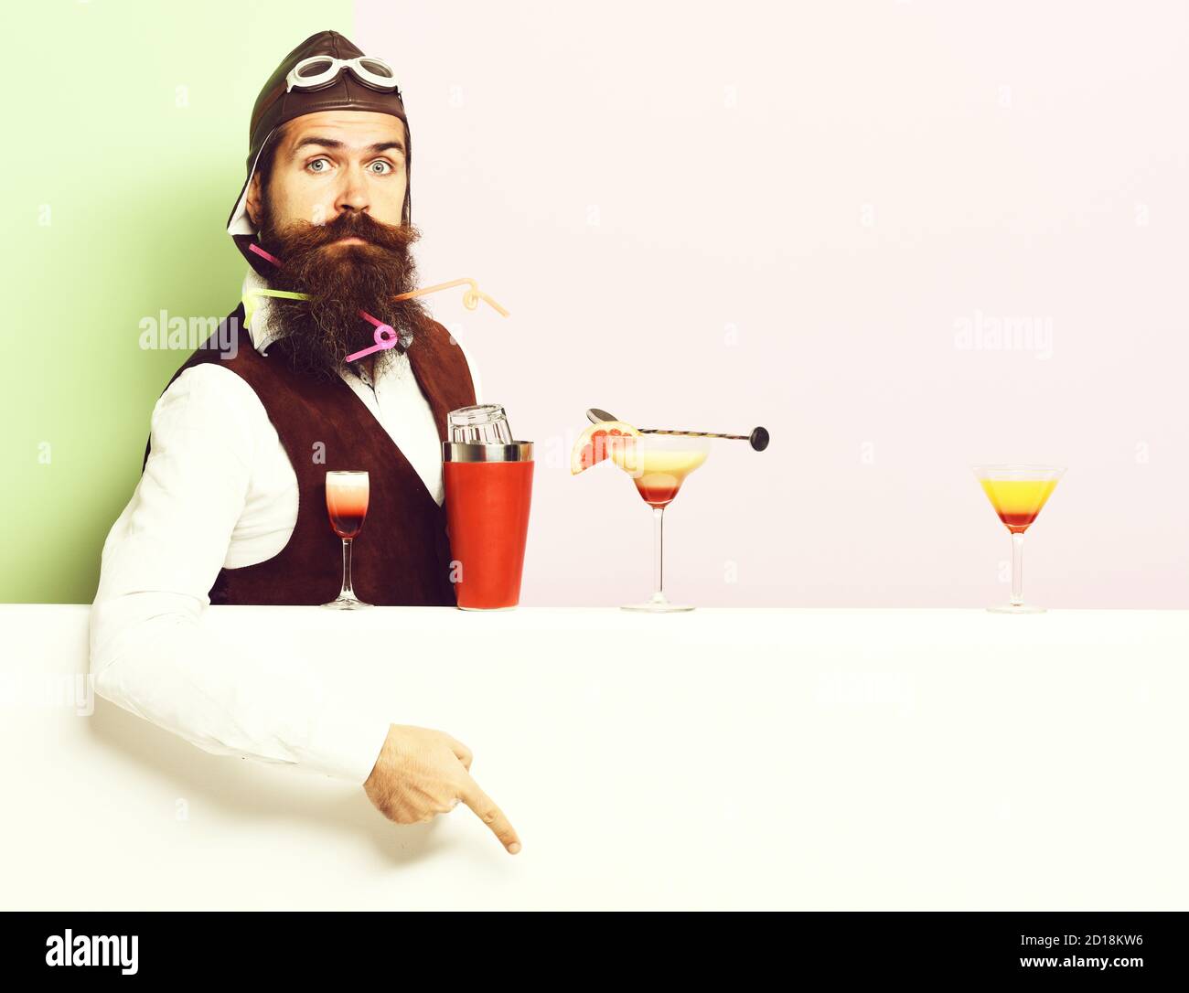 handsome bearded aviator or pilot with beard and mustache on surprised face with shaker alcoholic cocktails in vintage suede leather waistcoat with hand and glasses, on purple green studio background Stock Photo