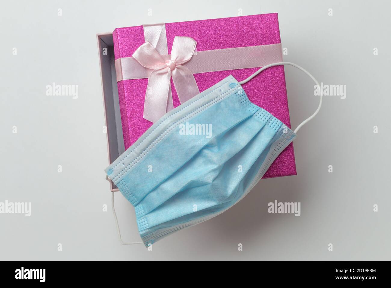 isolated small pink present box, open bright purple holiday gift box with a pink lace bow on top lying on a white background with a surgical blue face mask on top, idea of safe present, take care Stock Photo