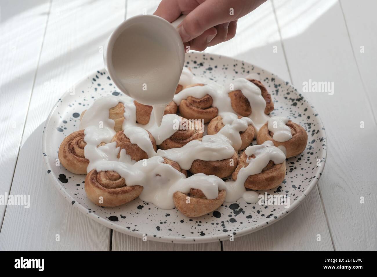 human hand pouring milk sauce or frosting on cinnamon swirls. selective focus. homemade cinnamon rolls with cream glaze. home cooking concept. Stock Photo