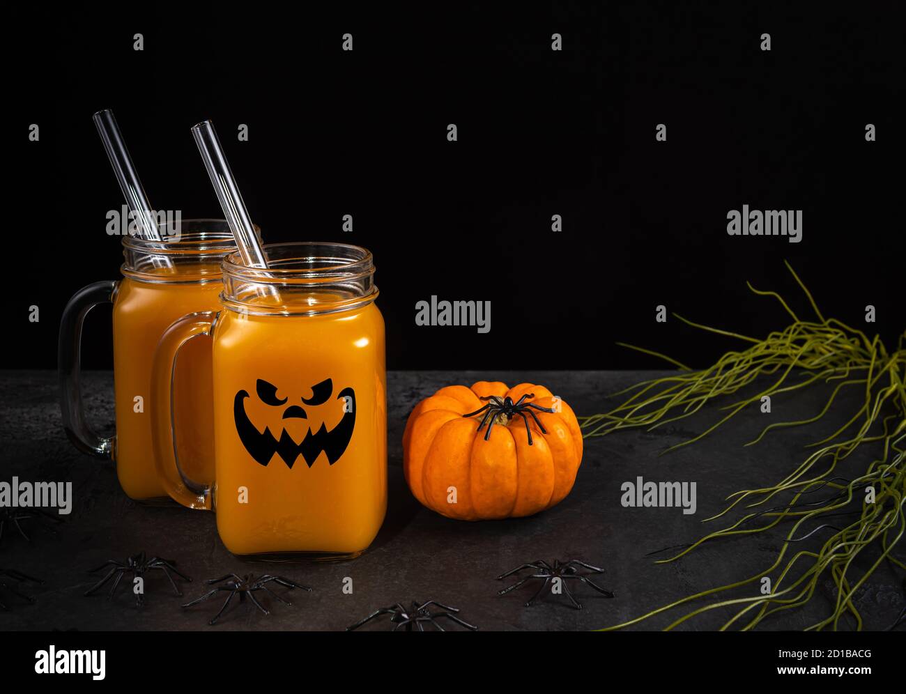 Glass jars with halloween face, pumpkin mocktail and glass straws on black textured table decorated with spiders, spooky plants and pumpkin. Horizonta Stock Photo