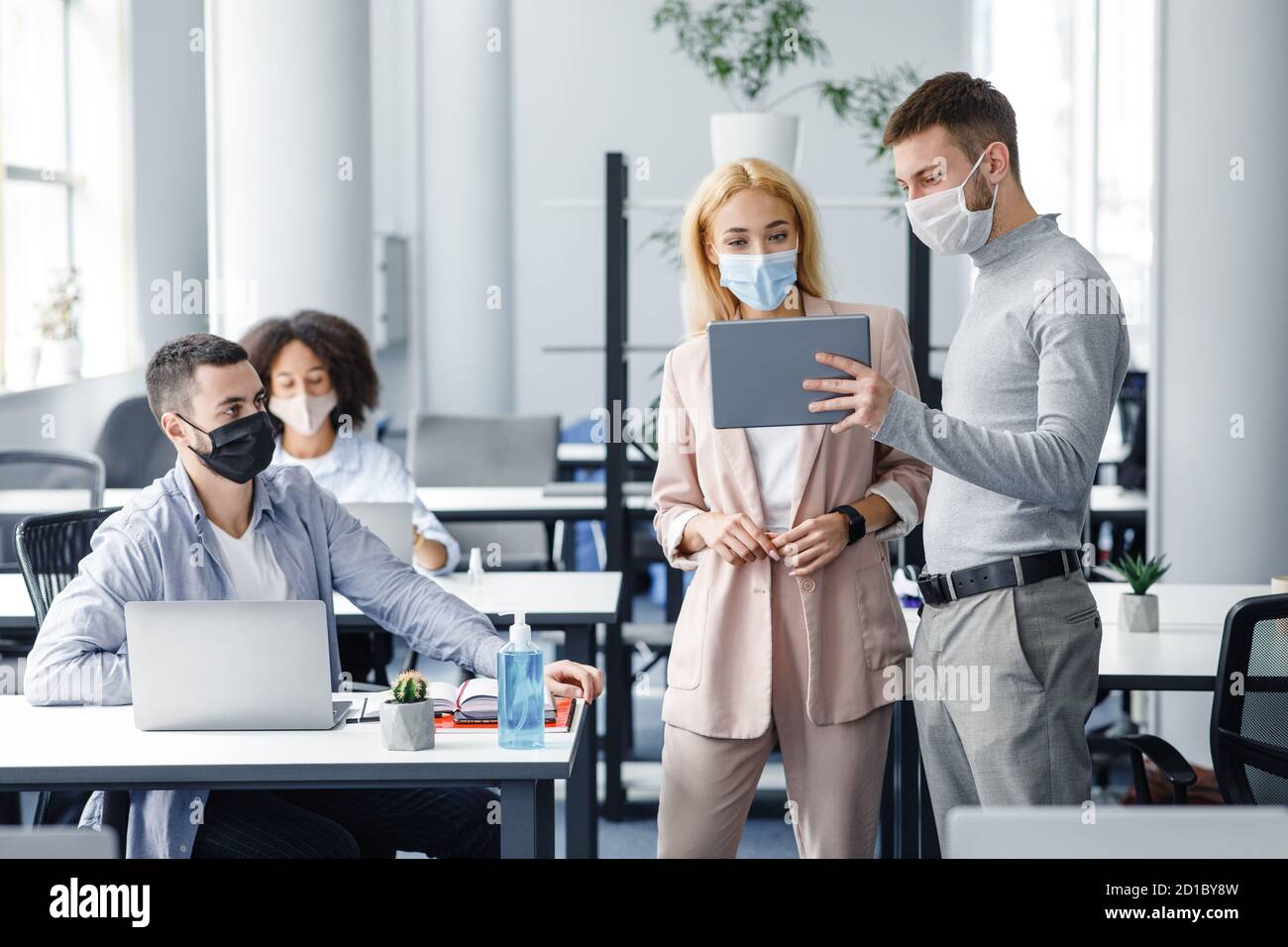 Teamwork in corporate company and social distance during coronavirus epidemic. Male manager in protective mask shows tablet to workers in interior Stock Photo