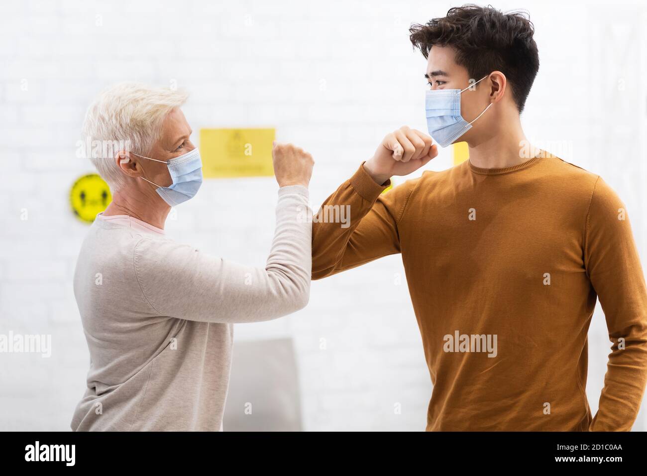 Guy And Senior Woman In Masks Bumping Elbows Meeting Indoors Stock Photo