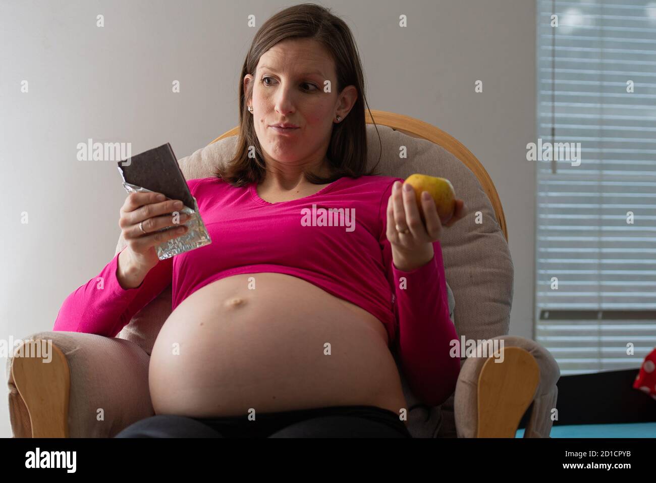 Young pregnant woman with big swollen belly deciding between a a healthy choice of an apple and an unhealthy chocolate. Stock Photo