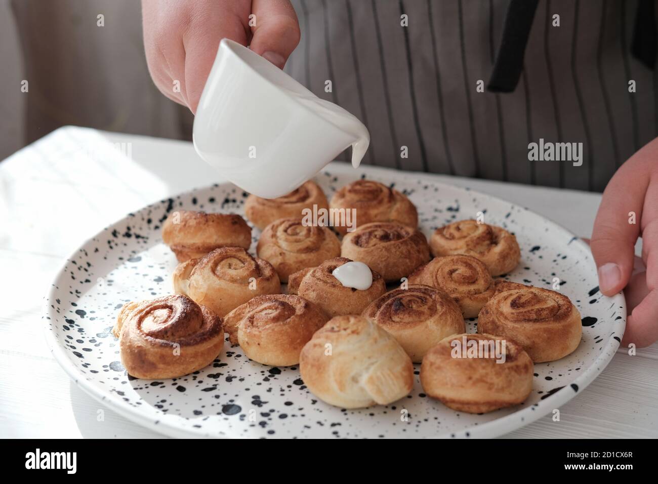 human hand pouring frosting on cinnamon swirls. selective focus. homemade cinnamon rolls with cream glaze. home cooking concept. close up view Stock Photo