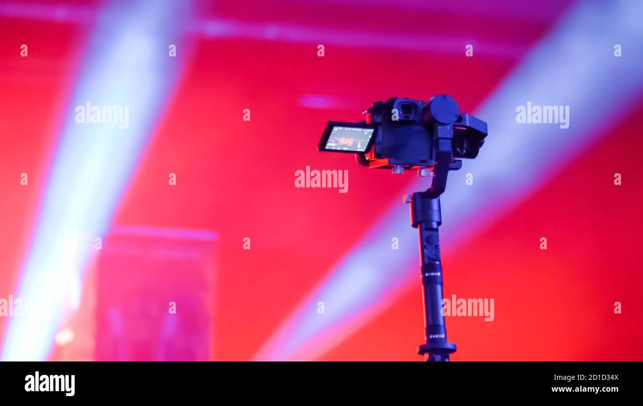 Digital mirrorless camera on handheld gimbal stabilizer taking photo or recording video of live music concert with red illumination. Photography Stock Photo