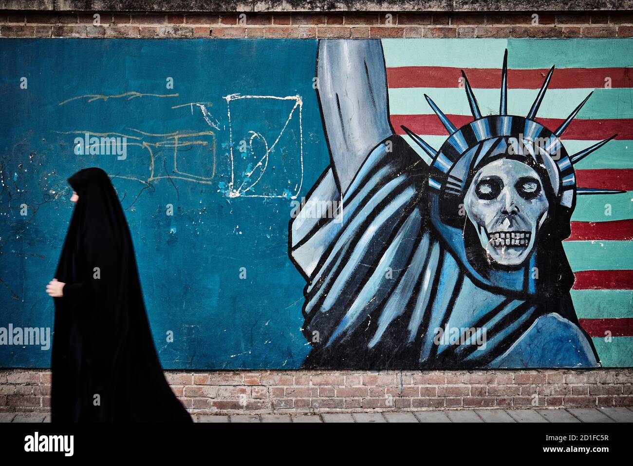 Islamic woman walks in front a wall of the old embassy of United States with a graffiti showing the USA flag with the Statue of Liberty with a skull Stock Photo