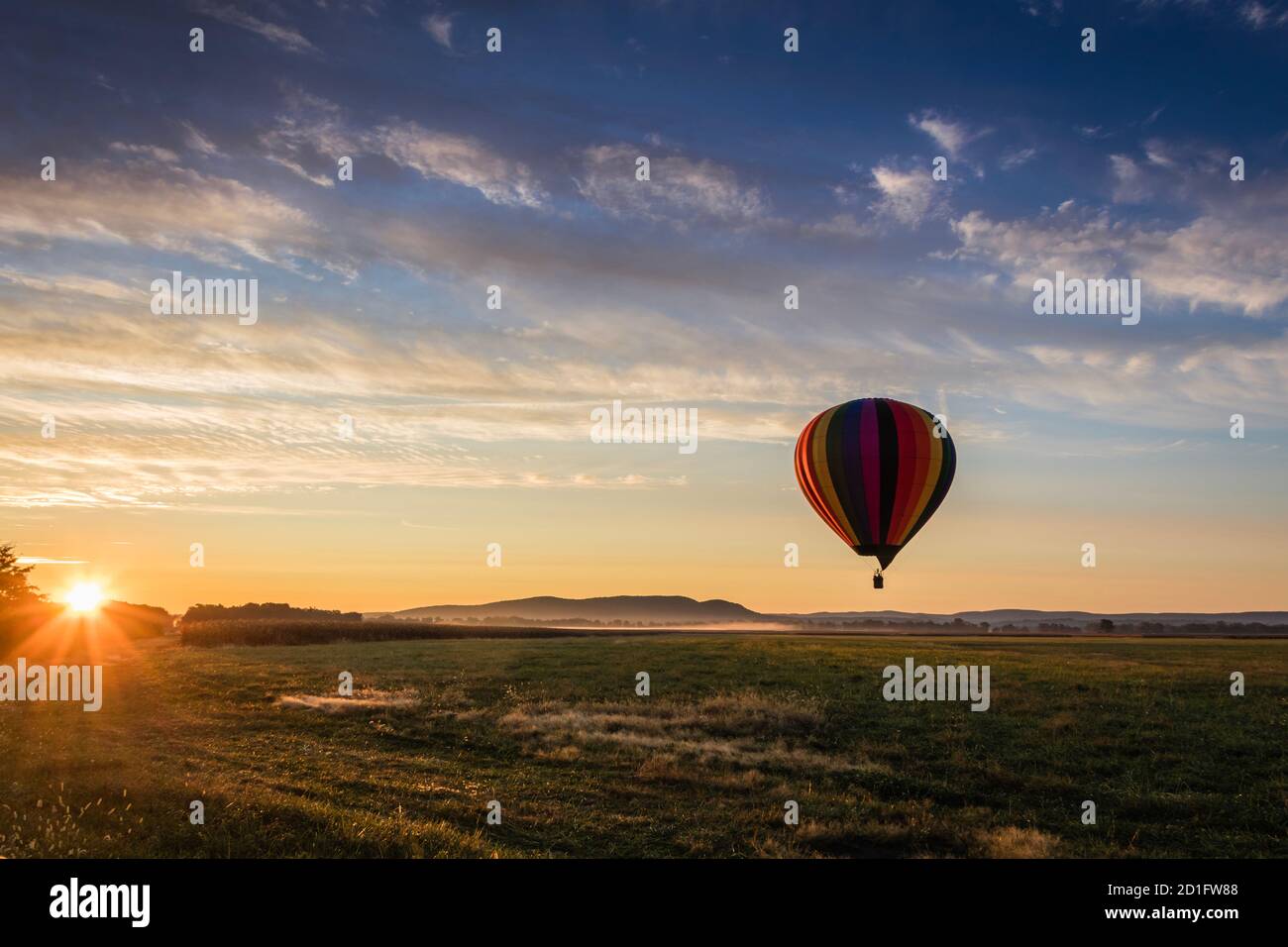 Hot Air Balloon in colorful rainbow stripes begins ascent over farm field as sun rises blue cloudy sky Stock Photo