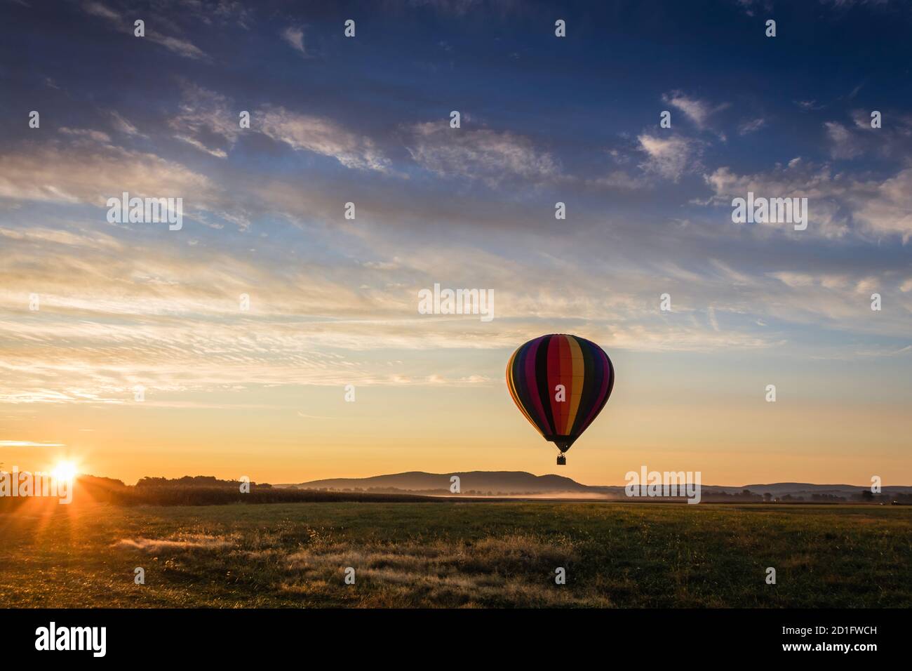 Hot Air Balloon in colorful rainbow stripes begins ascent over farm field as sun rises blue cloudy sky Stock Photo