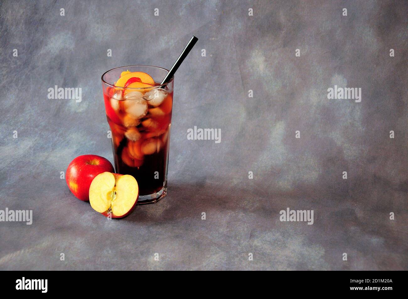A tall glass of red juice with ice, a stick and two ripe apples on a gray background. Close-up. Stock Photo