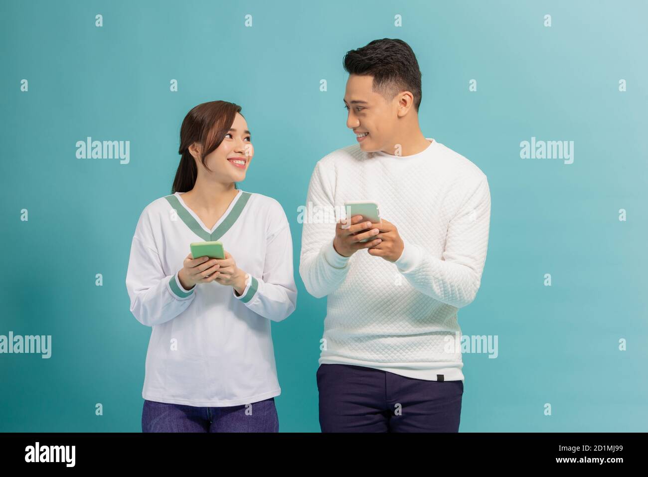 Cheerful young couple standing isolated over blue background, holding smart phones in casual wear Stock Photo