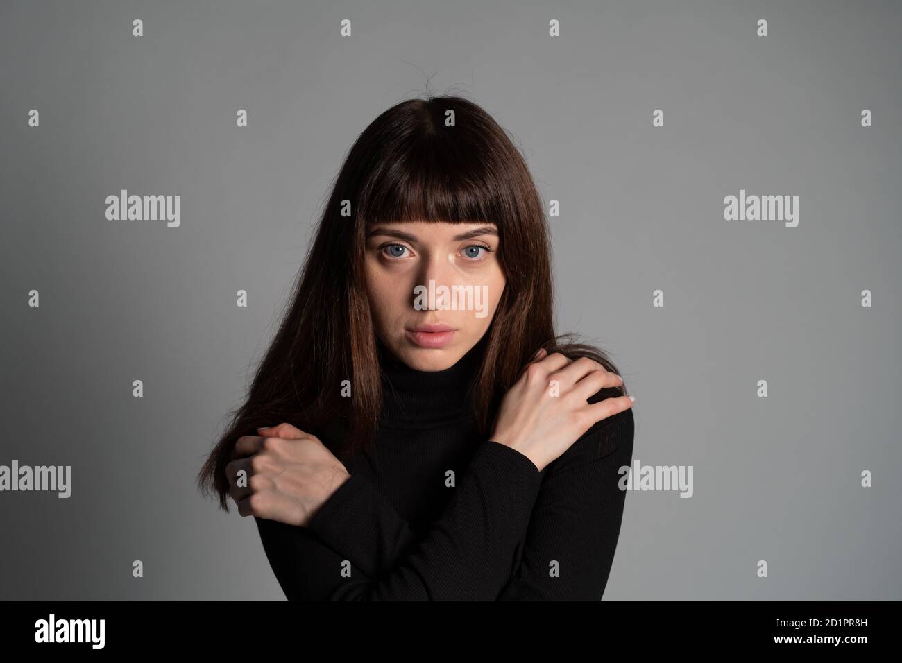 Close up studio portrait of a pretty brunette woman, wearing folded black polo-neck sweater, hands on shoulders, looking at camera, against a plain gr Stock Photo