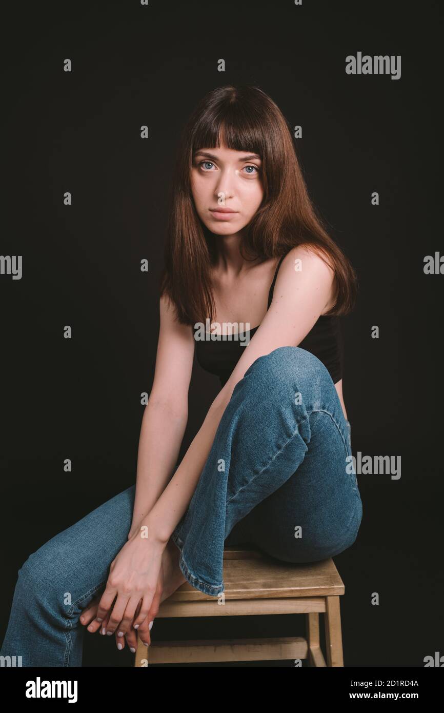 Studio portrait of a pretty barefoot brunette woman in black spaghetti strap top and jeans, looking at the camera, sitting against a plain black backg Stock Photo