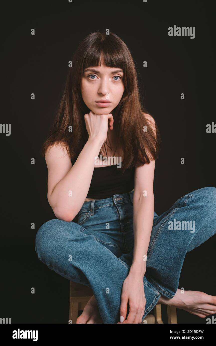 Studio portrait of a pretty barefoot brunette woman in black spaghetti strap top and jeans, looking at the camera, sitting, legs crossed, against a pl Stock Photo