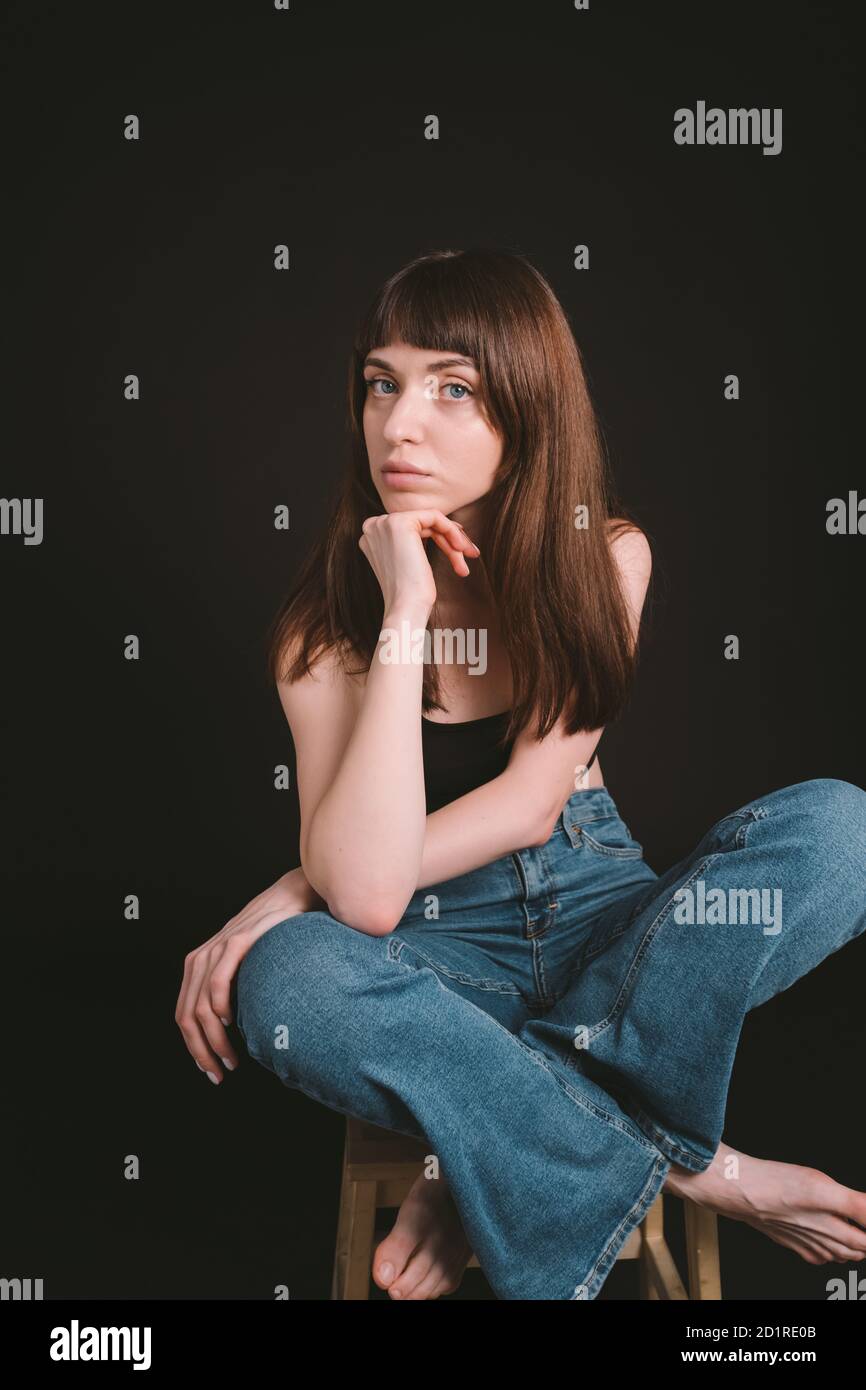 Studio portrait of a pretty brunette woman in black spaghetti strap top and jeans, looking at the camera, sitting, legs crossed, against a plain black Stock Photo