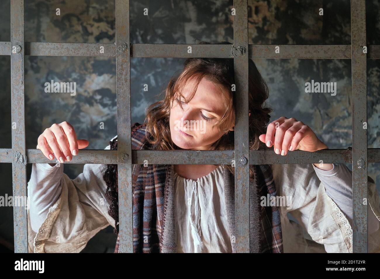A young woman imprisoned sadly looks out from behind bars in a medieval prison. Portrait of a criminal in vintage clothes at a Halloween party Stock Photo