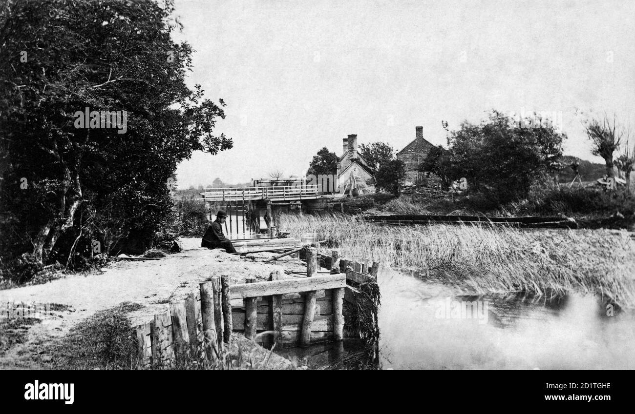 ST JOHN'S LOCK, Lechlade, Gloucestershire. The weir and lock keeper's house at St John's Lock on the River Thames. A man sits on the riverbank and contemplates the reeds. (Published in Taunt's 'New Map of the River Thames', 5th edition, 1886-7). Photographed between 1860 and 1887 by Henry Taunt. Stock Photo
