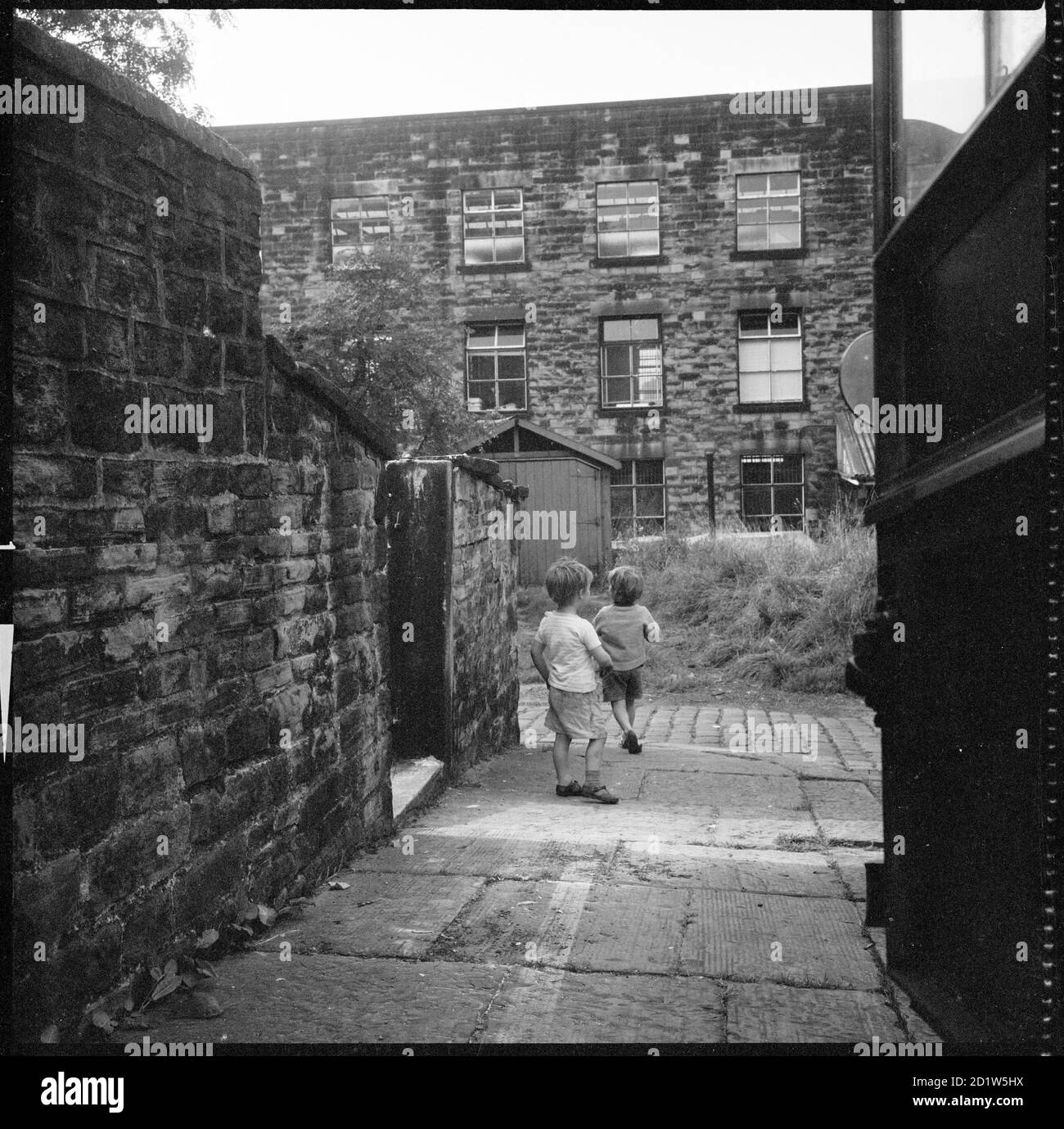 Holker Street Mill seen from the lane running between 48 Burnley Road and Bethel Church, with two children playing in the lane, Colne, Pendle, Lancashire, UK. Stock Photo