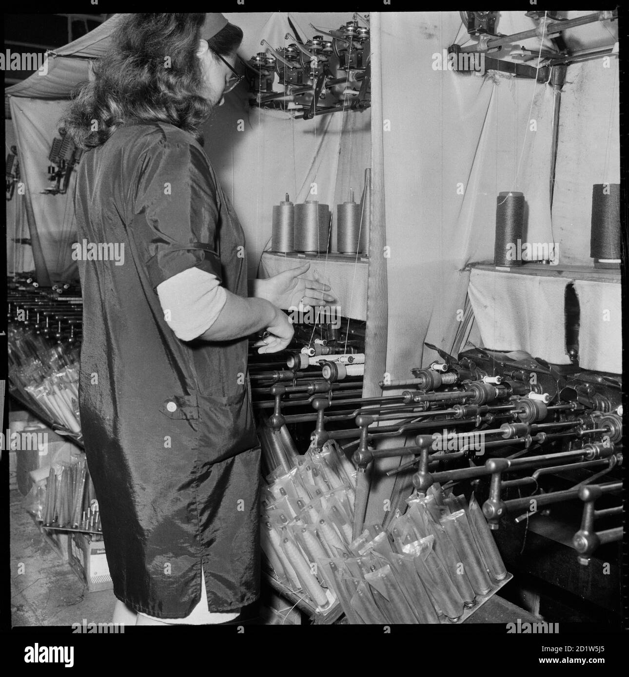 A woman operating a cop winding frame with wound cops can be seen hanging from the machine. Stock Photo