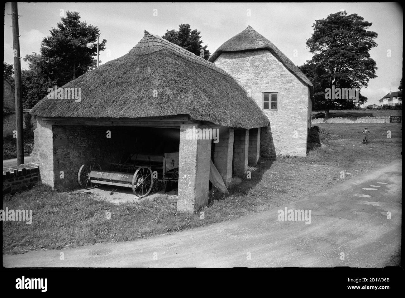 Exterior view of a single-storey thatched barn attached to a stone building with thatched roof and housing a wooden vehicle, likely a cart or trap, set within a low-walled yard with a man stood nearby and a large house in the background. Stock Photo
