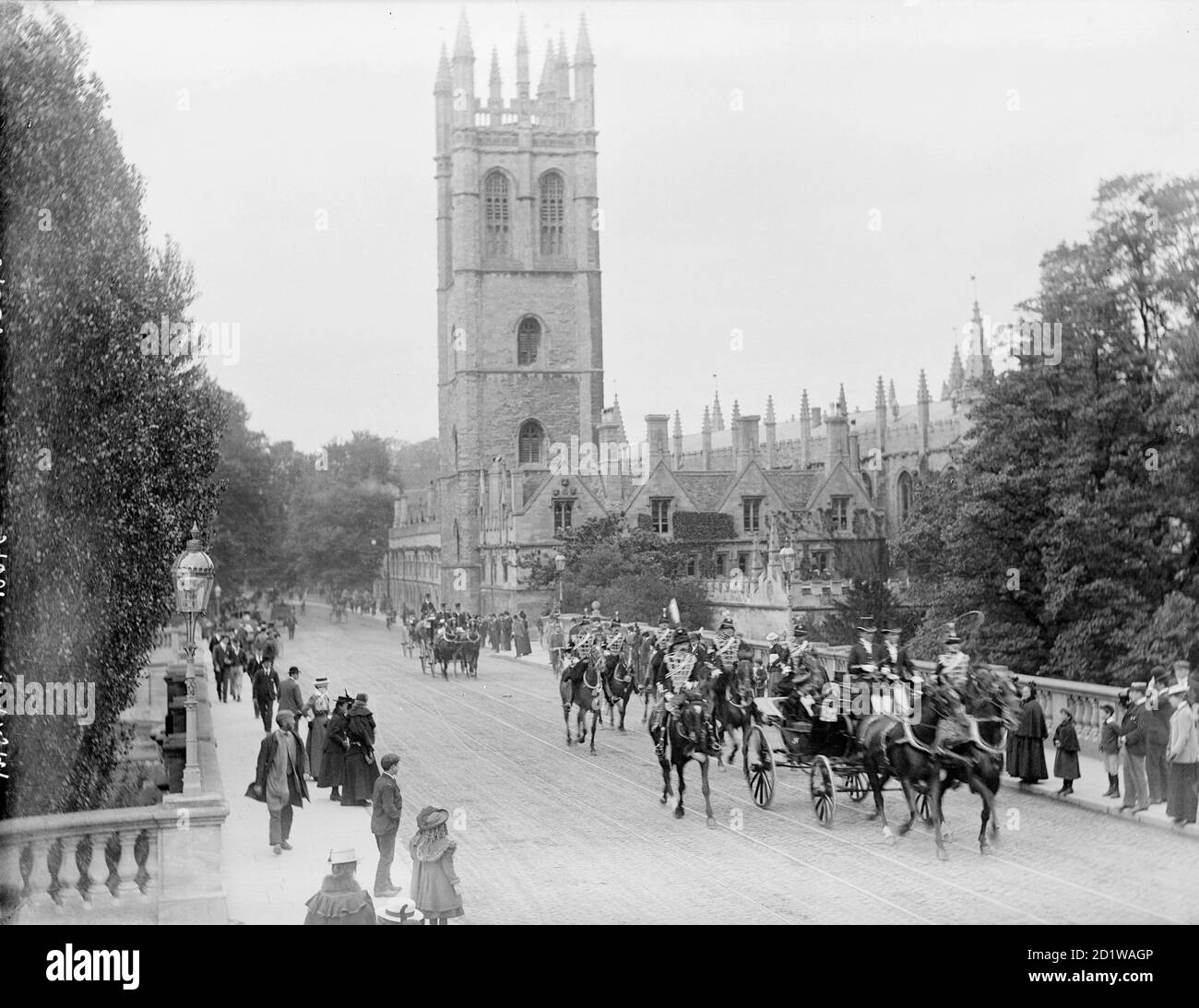 High Street, Oxford, Oxfordshire. A procession of horse drawn carriages and guards passing in front of Magdalen College, watched by onlookers from the pavement. Stock Photo