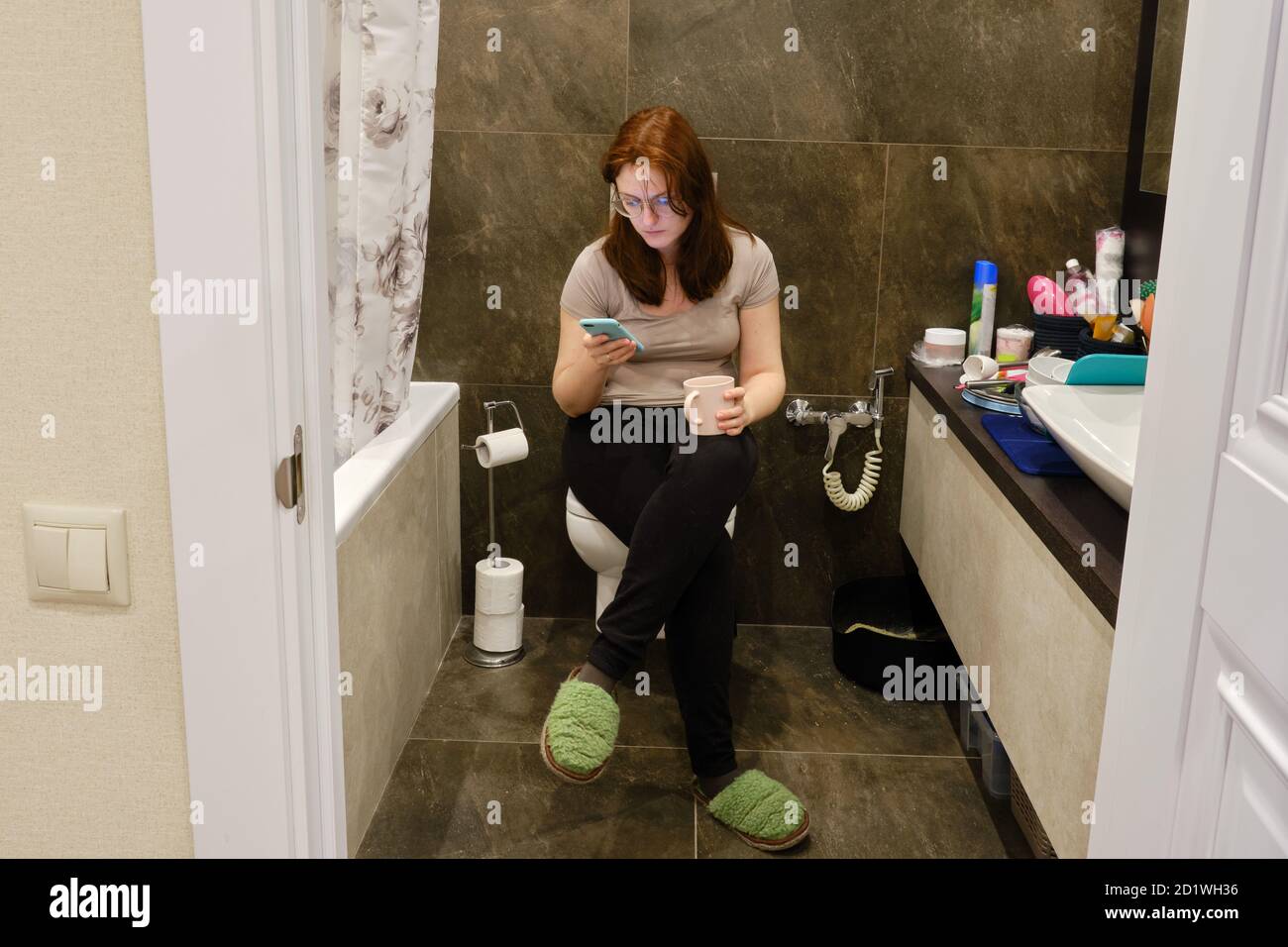 A young woman looks at her phone while sitting on the toilet in the bathroom. Lifestyle photos and casual life Stock Photo