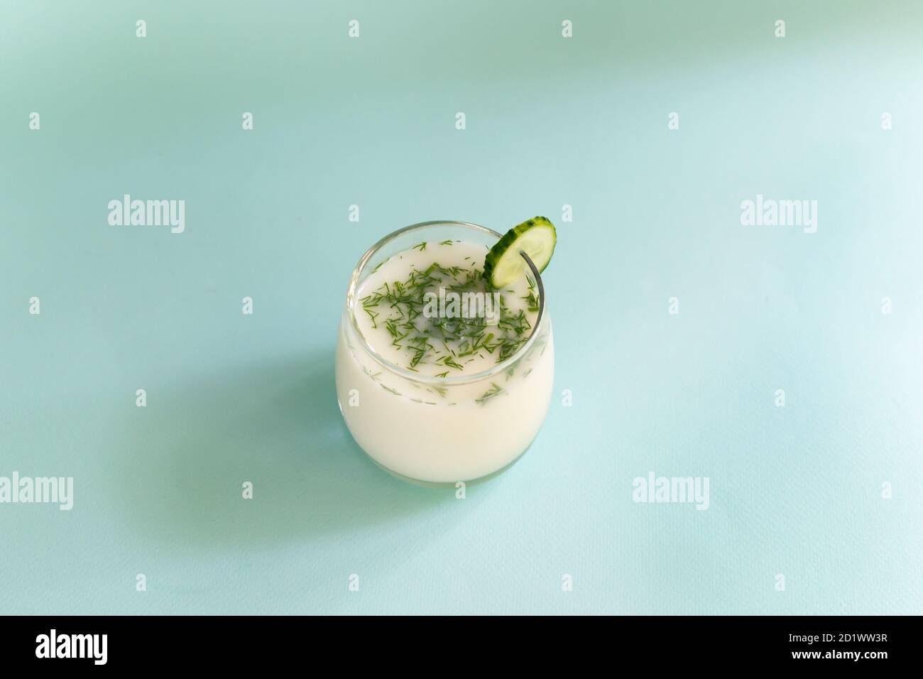 Ayran in a glass glass with dill and a slice of cucumber on a blue background. Fermented product concept. Copy space. Stock Photo