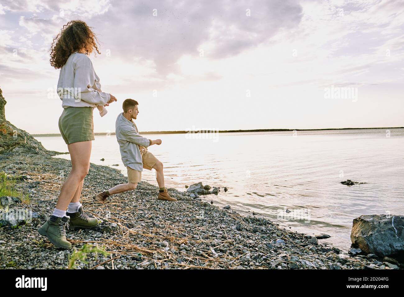 Young man standing with girlfriend at lake shore and throwing stones in water while his girlfriend looking at him Stock Photo