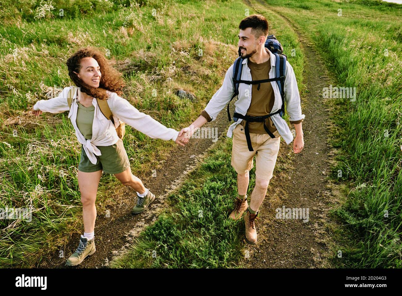 Above view of young couple of hikers with backpacks holding hands and walking over field with wild grass Stock Photo