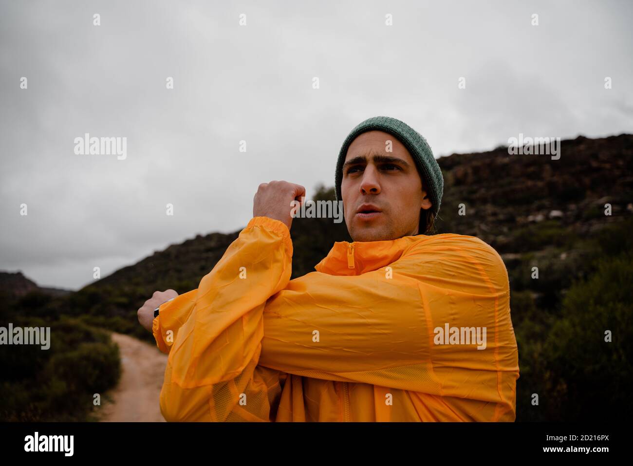 Young attractive male athlete focused on stretching arms before long distance run up mountain trail in cloudy weather Stock Photo