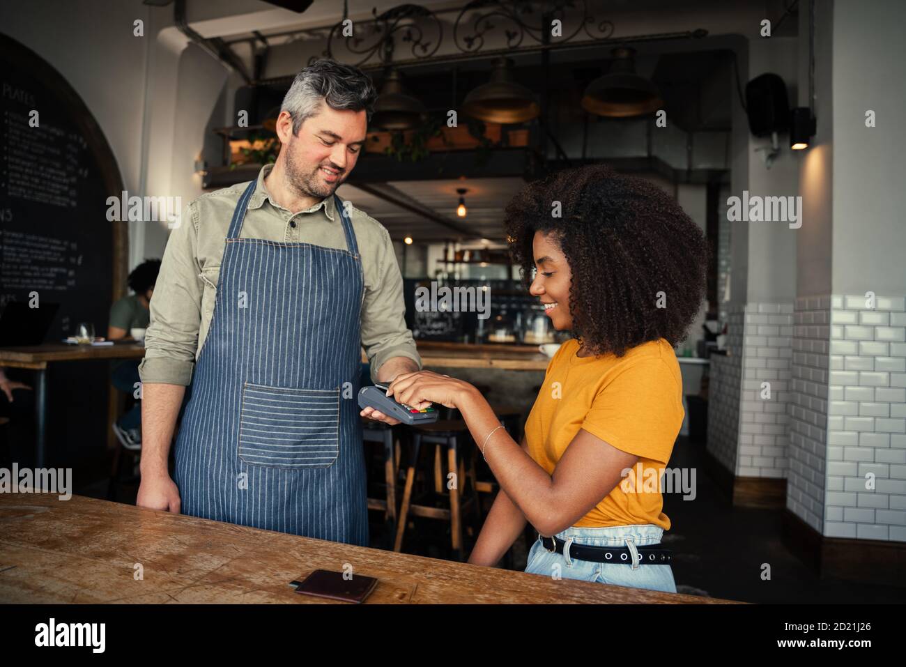 Beautiful ethnic woman tapping card making payment for coffee to handsome waiter in trendy cafe. Stock Photo