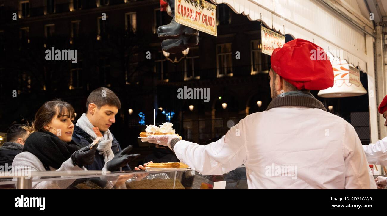 PARIS, FRANCE - JANUARY 6, 2019:  Young woman buying delicious waffle with whipped cream at Christmas market stall selling fresh waffles, crepes, hot Stock Photo