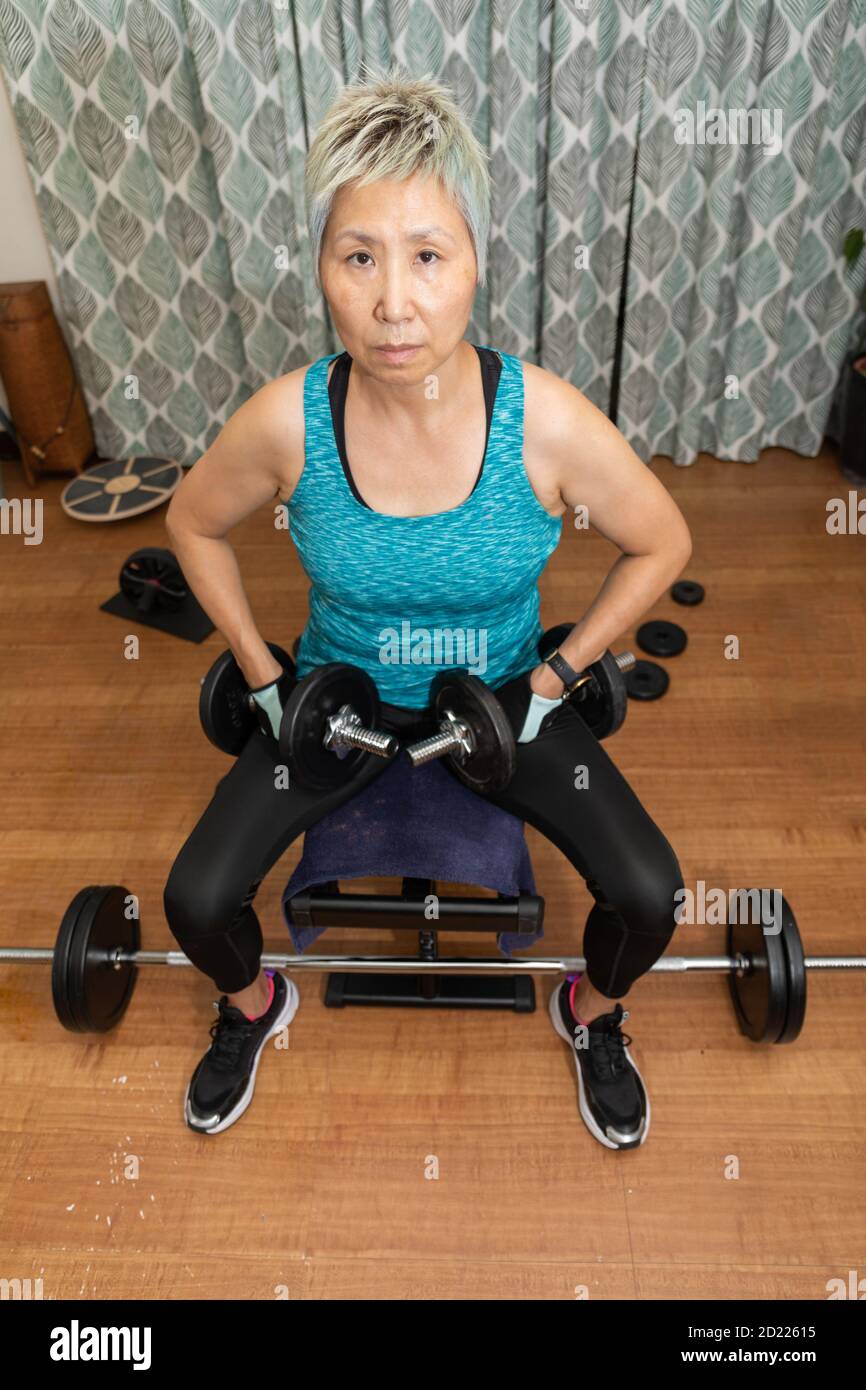 Fit mature Taiwanese woman of Chinese ethnicity exercises alone in her home to avoid crowds during the covid-19 pandemic. Stock Photo