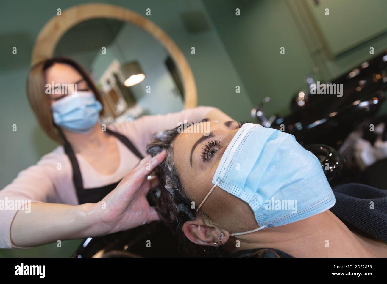 Female hairdresser wearing face mask washing hair of female customer at hair salon Stock Photo