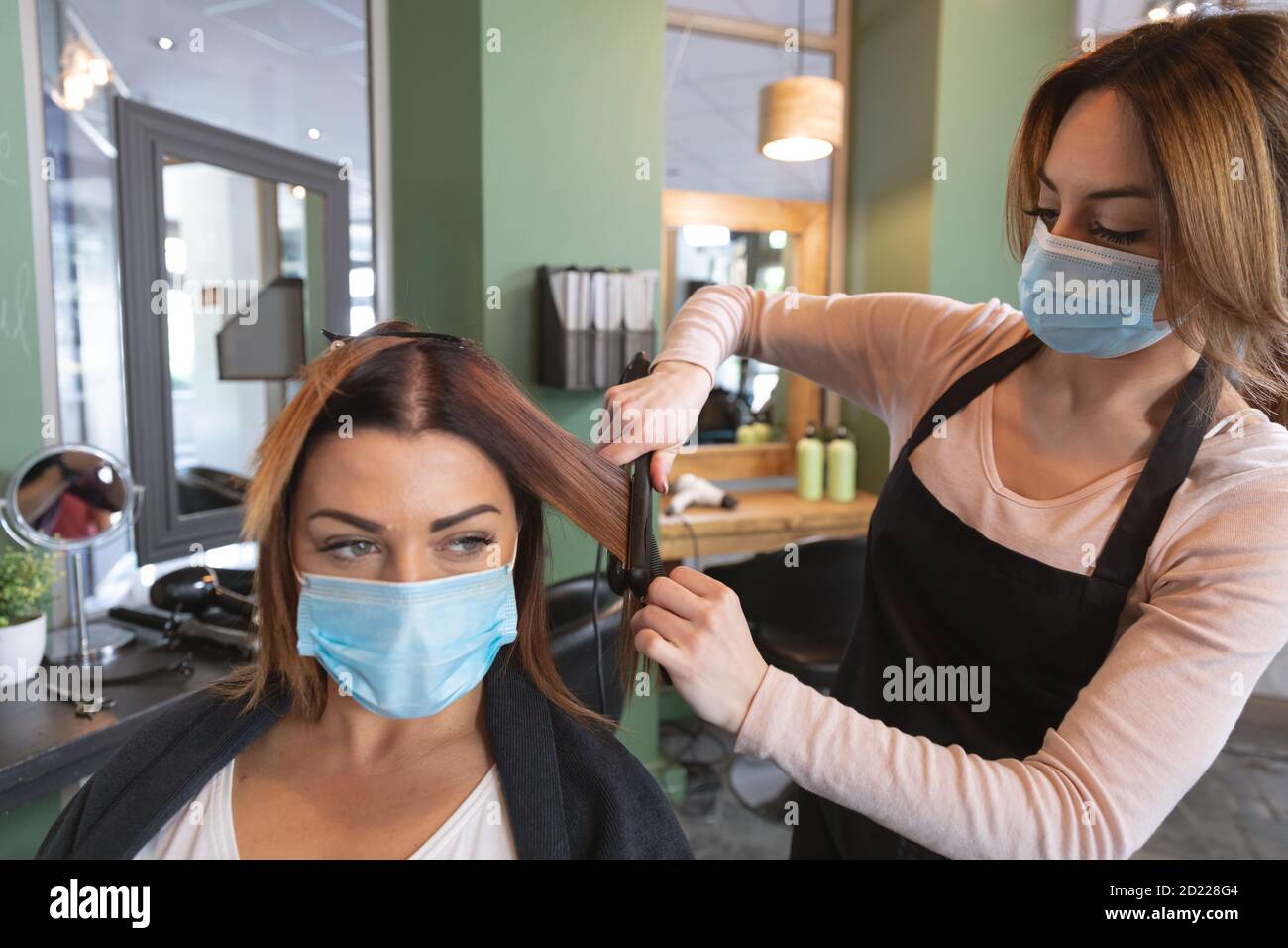 Female hairdresser wearing face mask straightening hair of female